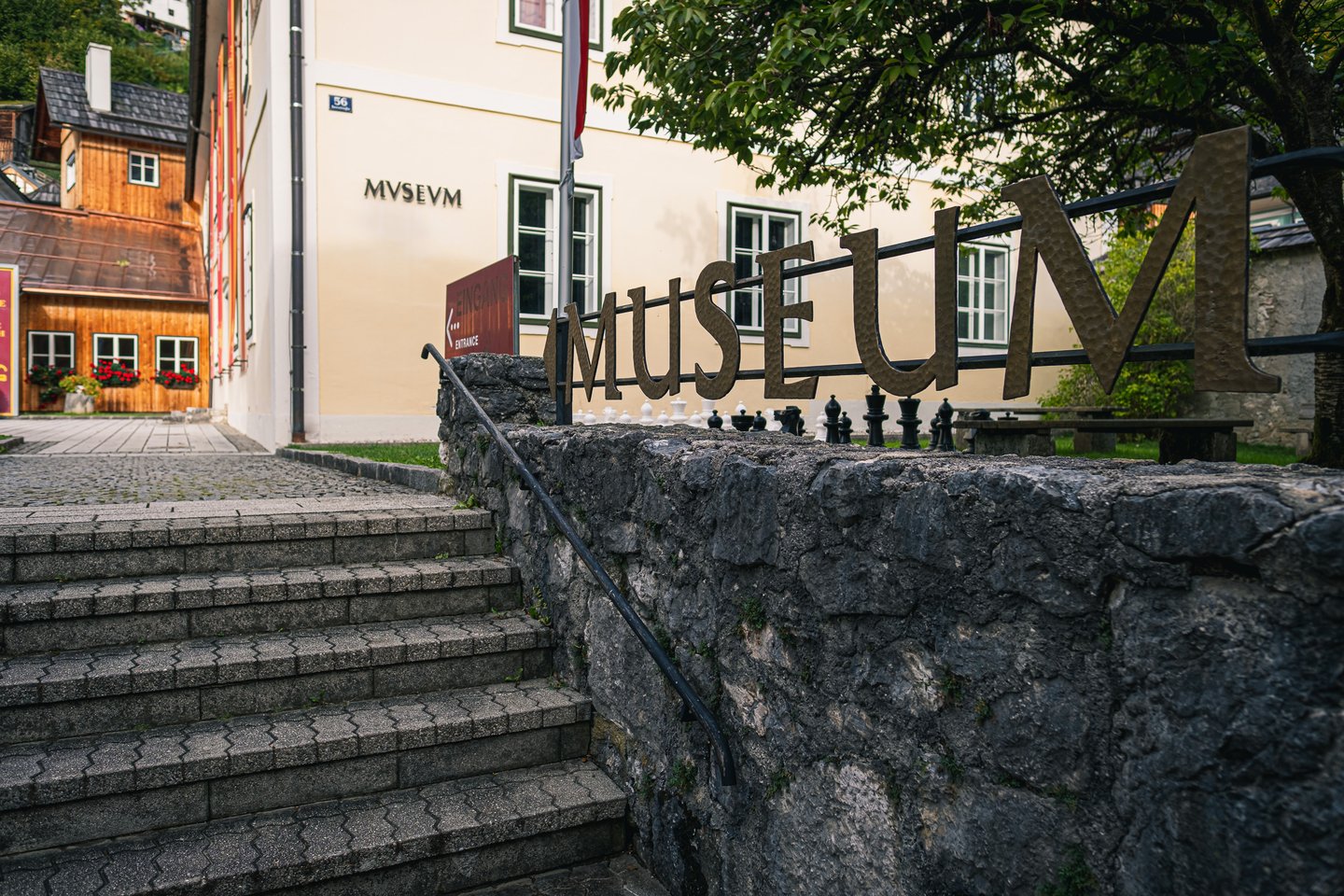 Hammered metal letters spelling MUSEUM run along a railing on a low stone wall outside the Hallstatt Museum, with a giant outdoor chess set behind them and the cream museum building with its MVSEVM lettering and entrance arrow beyond.