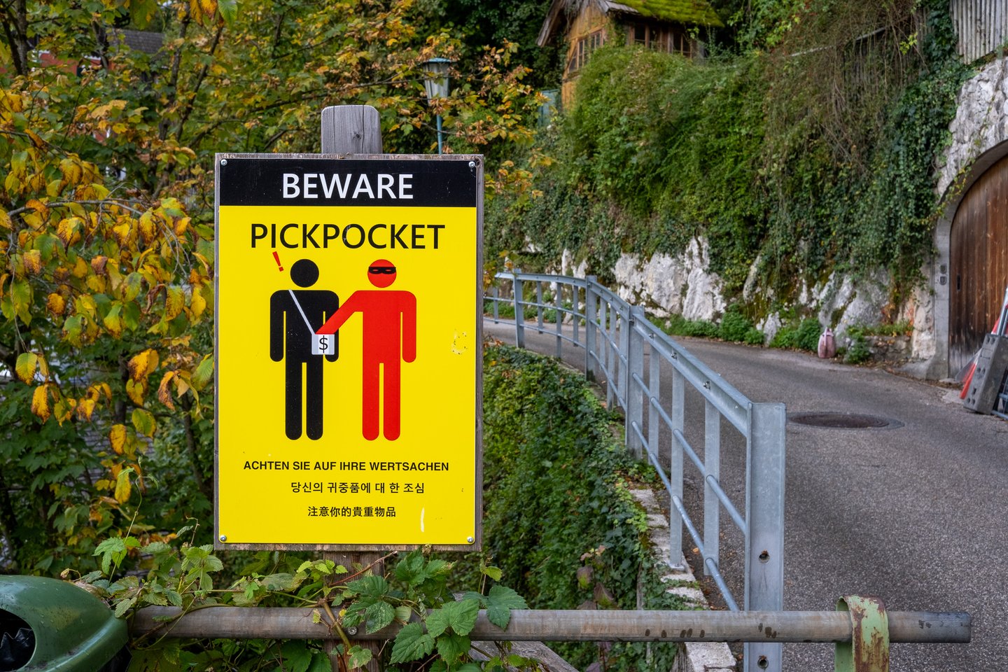 A yellow BEWARE PICKPOCKET warning sign in German, Korean and Chinese stands beside a narrow lane in Hallstatt, with a guard rail, ivy-covered rock and autumn foliage behind it.