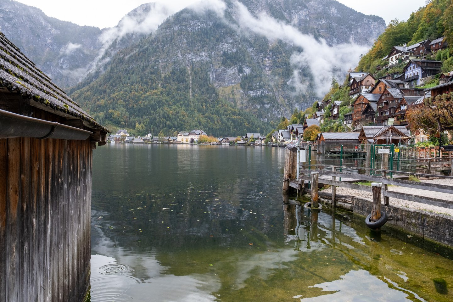 The Hallstaetter See seen from the village shore, with a weathered boathouse wall on the left, mooring posts and a fenced jetty on the right, and cloud hanging halfway down the forested mountain across the water.