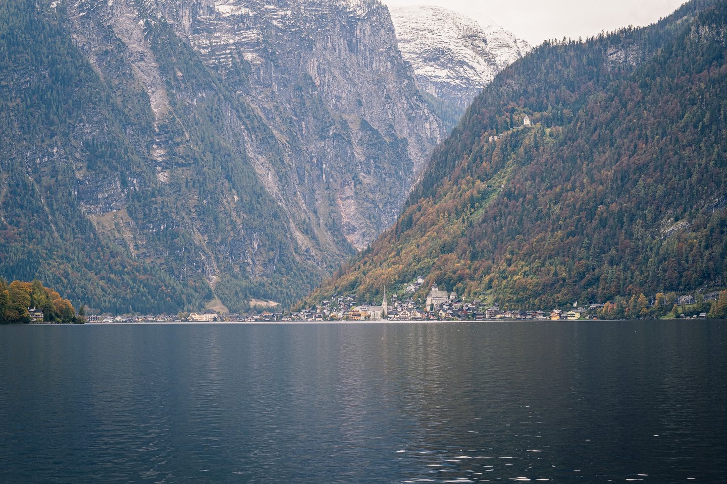 A wide view up the Hallstaetter See to the village at the foot of the mountains, with the first snow of the season on the grey peaks and the wooded slopes turning rust and gold.