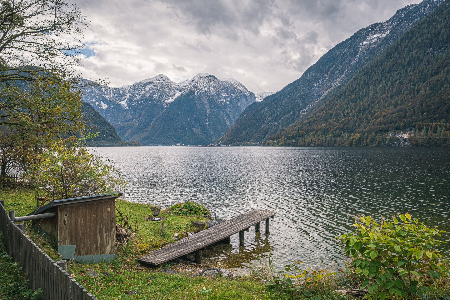 A weathered plank jetty and a small wooden shed sit on the grassy shore of the Hallstaetter See, looking up the lake to peaks under the season's first snow.