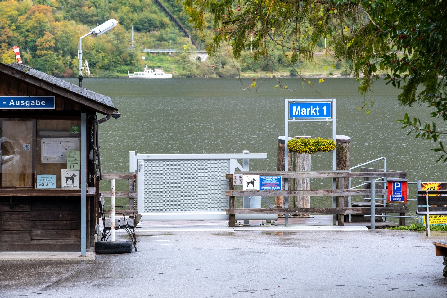 The Markt 1 landing stage on the Hallstaetter See, with a blue enamel Markt 1 sign above a flower box on a mooring post, a closed gangway gate, a wooden ticket hut carrying timetables and a small motorboat out on the grey water.