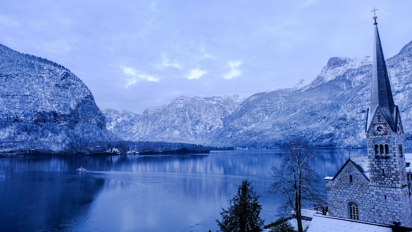 At winter dusk the Hallstatter See lies flat and blue between snow-dusted mountain walls, a small ferry crossing at left and the stone spire of the Hallstatt evangelical church at right.