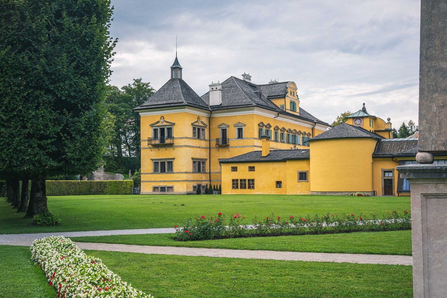 Yellow baroque Hellbrunn Palace with garden and pathway under overcast sky in Salzburg