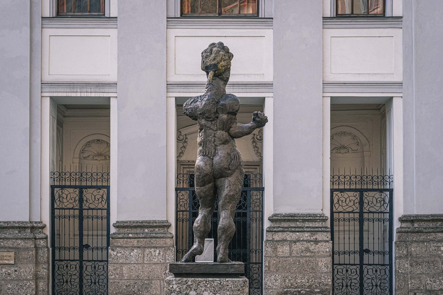 Homage to Mozart sculpture framed by the columned facade and iron gates of Markuskirche in Salzburg.