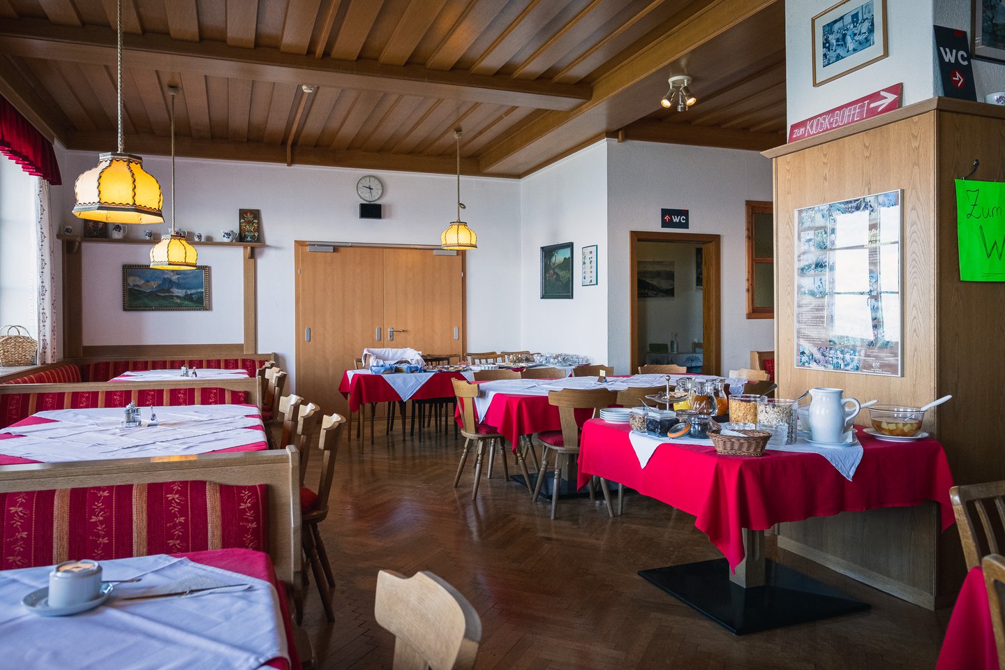 A mountain inn dining room with parquet floor, a beamed wooden ceiling and yellow glass pendant lamps, red draped tables and a hand painted sign pointing to the kiosk and buffet.