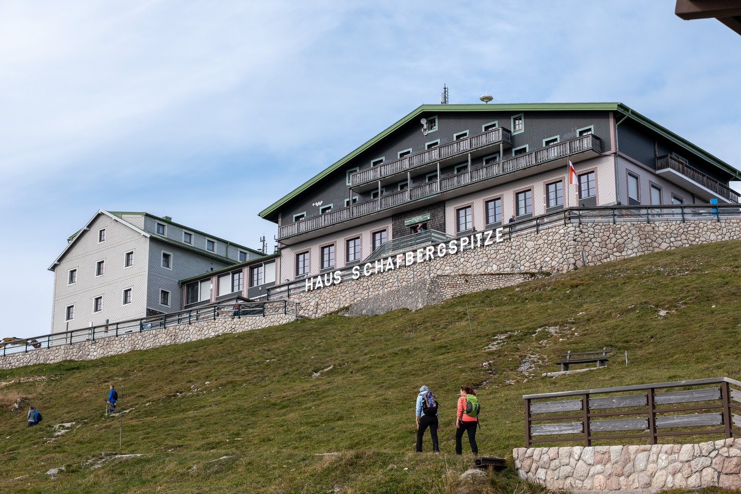 Looking up from the summit meadow, the whole Hotel Schafbergspitze rises above a stone retaining wall lettered HAUS SCHAFBERGSPITZE, its tiers of wooden balconies catching the light as two hikers climb the slope.