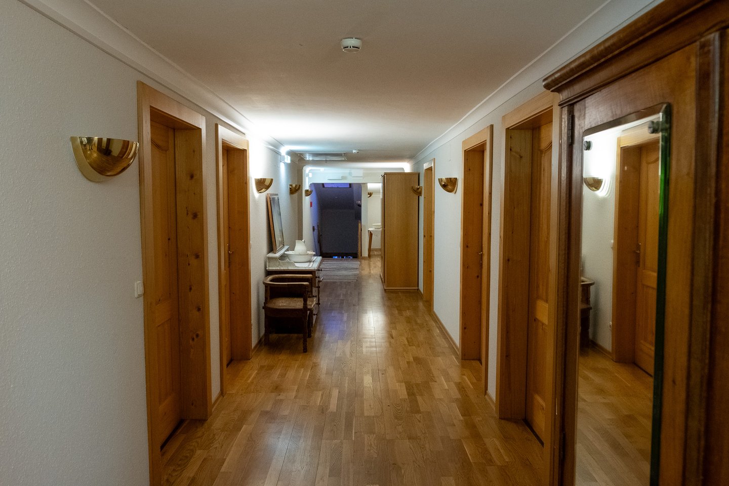 A quiet guest corridor with pale pine doors, brass half-shell wall lights and an oak plank floor, an antique washstand and chair at the far end, photographed before dawn.