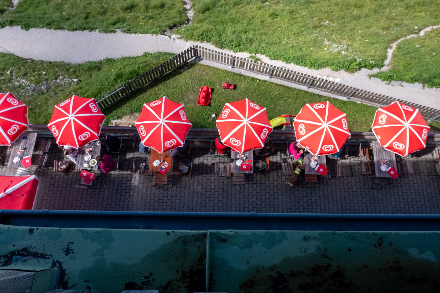 Looking straight down from an upper window onto a row of red and white parasols shading wooden benches on the paved summit terrace, with a picket fence and green slope beyond.