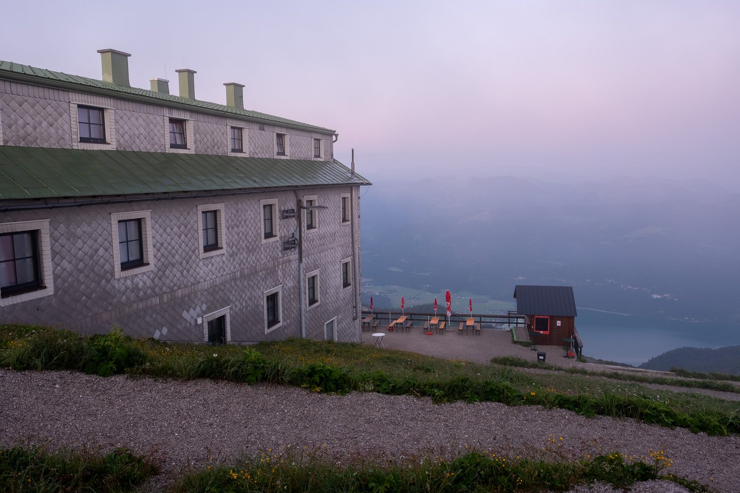 The long shingled flank and green metal roof of the Hotel Schafbergspitze at dawn, with its terrace of furled red umbrellas and a small kiosk hut looking down to the Wolfgangsee.