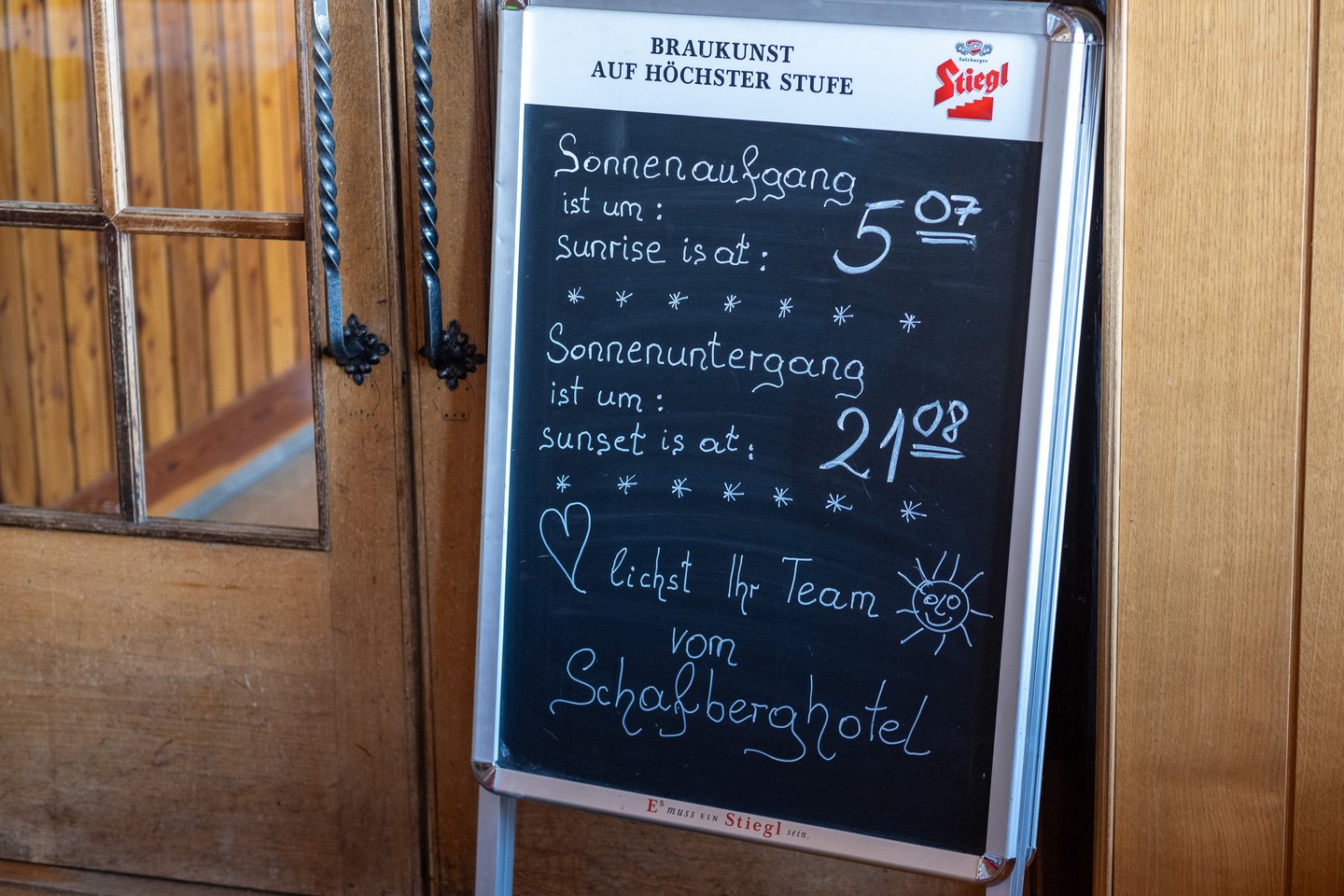 A chalkboard by the dining room doors of the summit hotel gives the day's sunrise as 5:07 and sunset as 21:08, signed with a drawn sun and a heart by the team of the Schafberghotel.