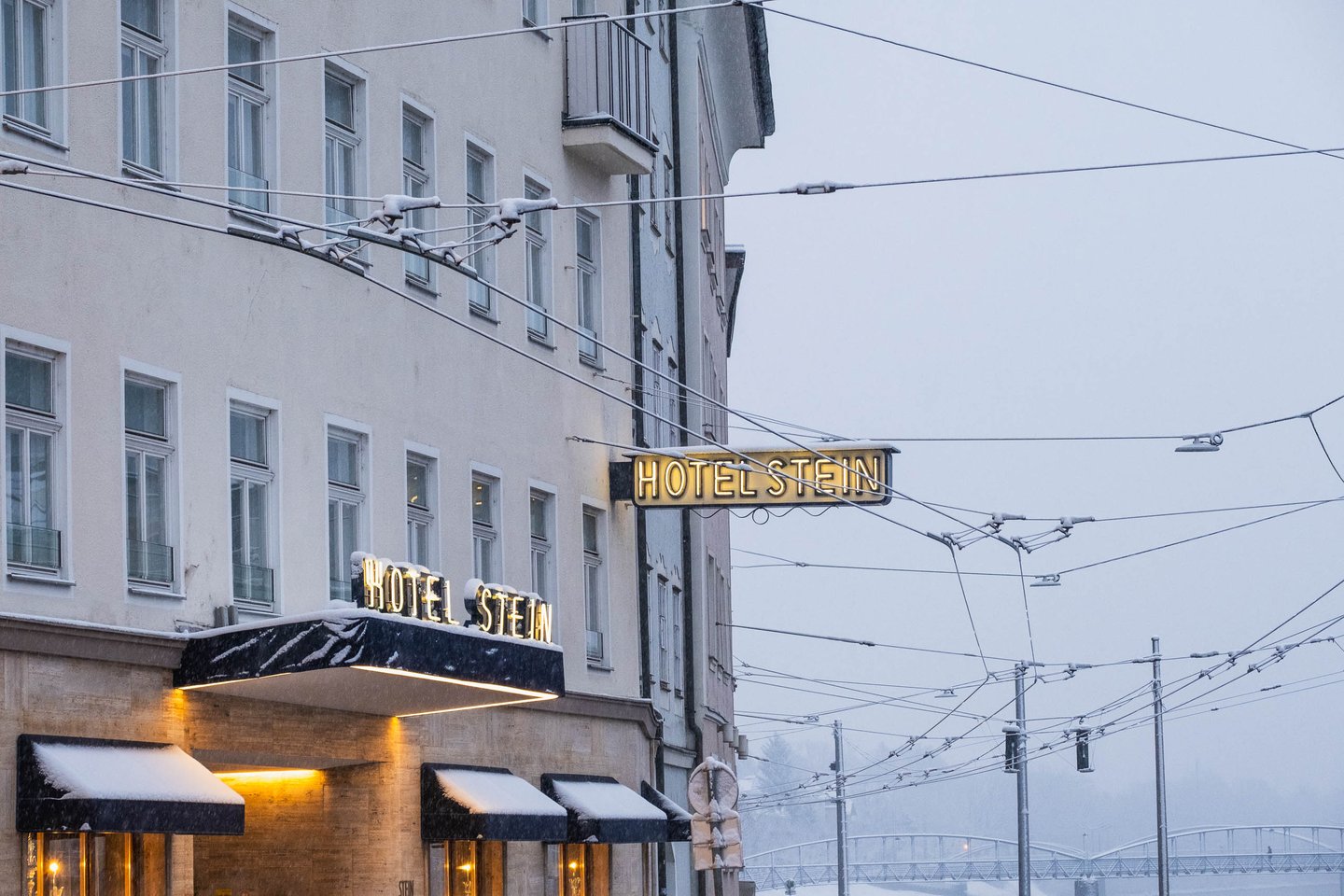 Hotel Stein facade and illuminated sign during snowfall near the Staatsbruecke tram lines in Salzburg.