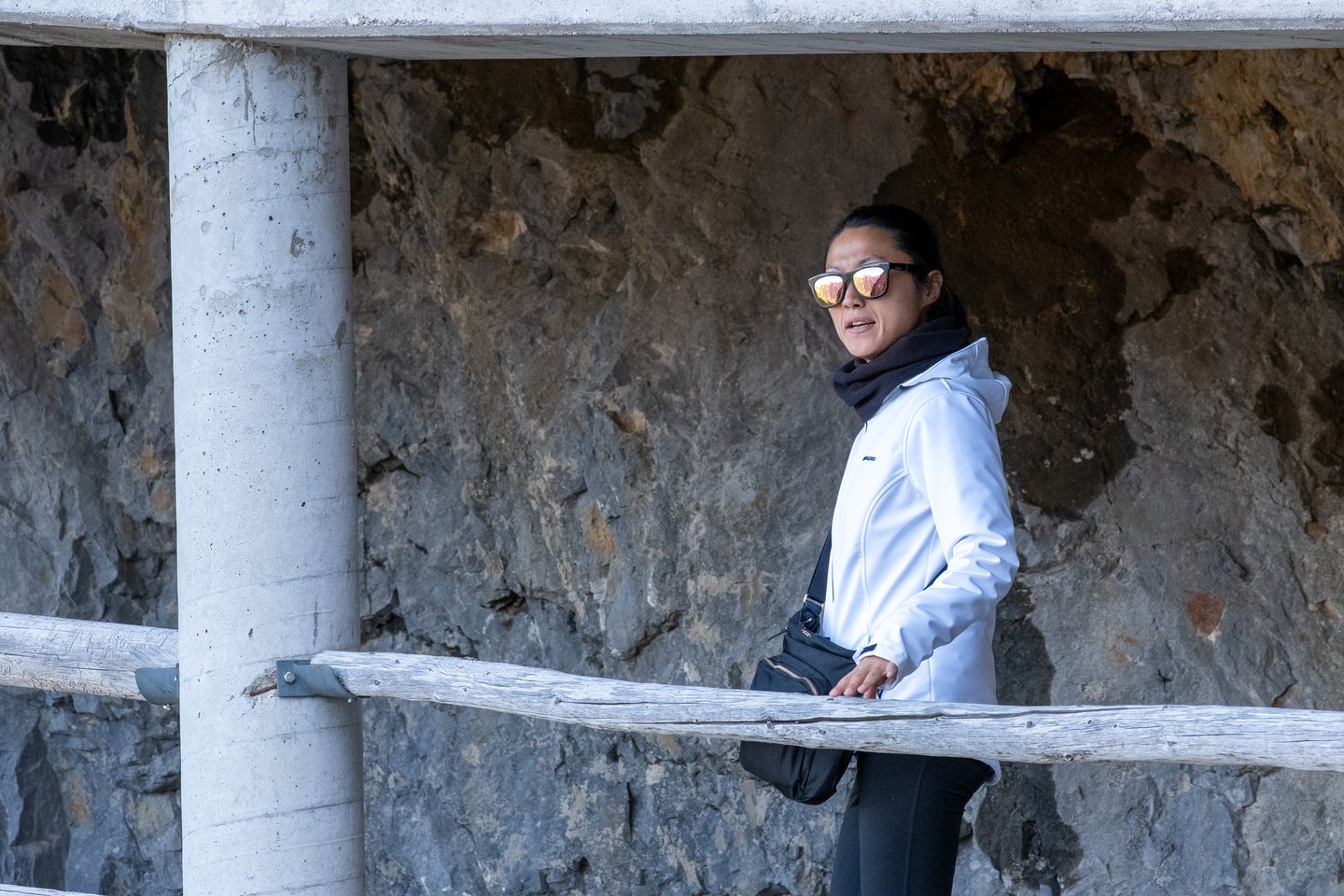 Hyunok Jang in a white jacket and mirrored sunglasses stands at a wooden railing under the concrete gallery near the Eisriesenwelt cave entrance.