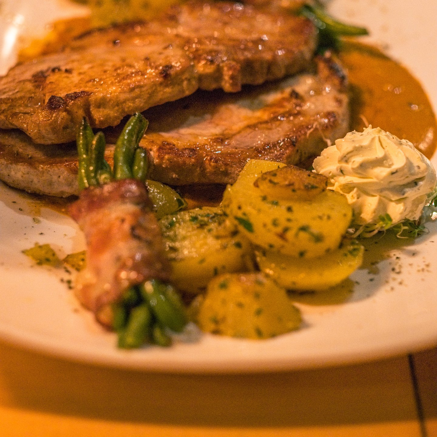 Two pan-fried pork cutlets on a white plate with green beans, parsley potatoes, a bacon roll and a quenelle of herb butter, in warm restaurant light.