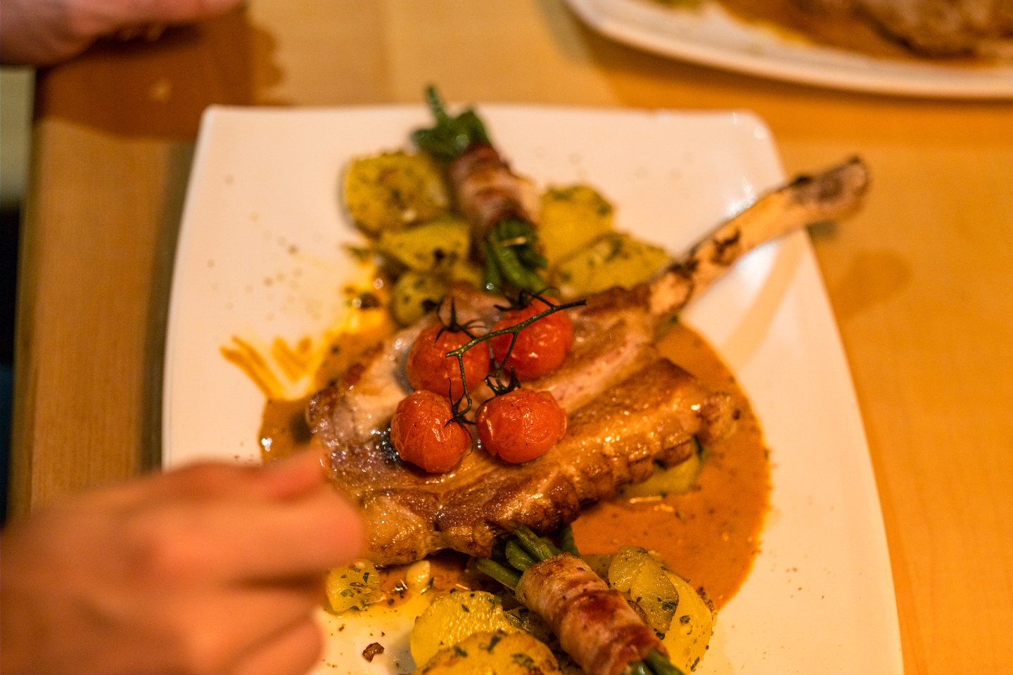 A bone-in pork chop topped with a truss of roasted cherry tomatoes sits in a paprika-coloured sauce beside parsley potatoes and bacon-wrapped green beans on an angular white plate.