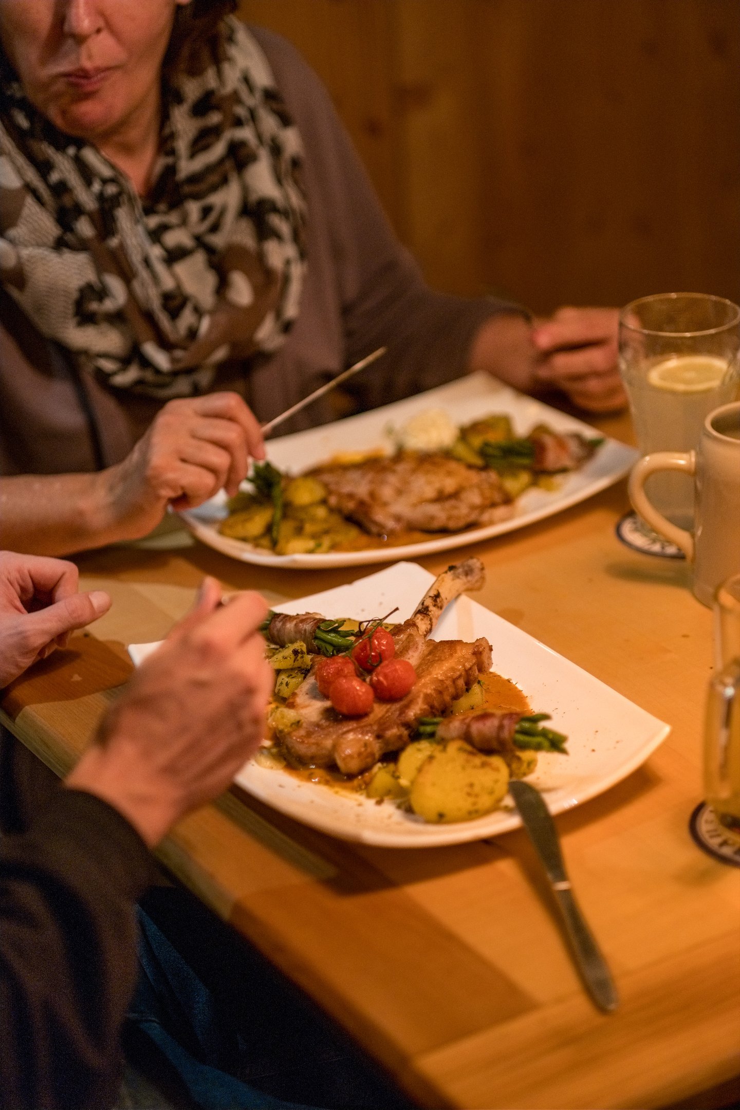Two diners at a wooden restaurant table, one plate of pork chop crowned with roasted cherry tomatoes in the foreground and another being eaten across the table, with ceramic beer mugs and a lemon-topped glass at the edge of the frame.