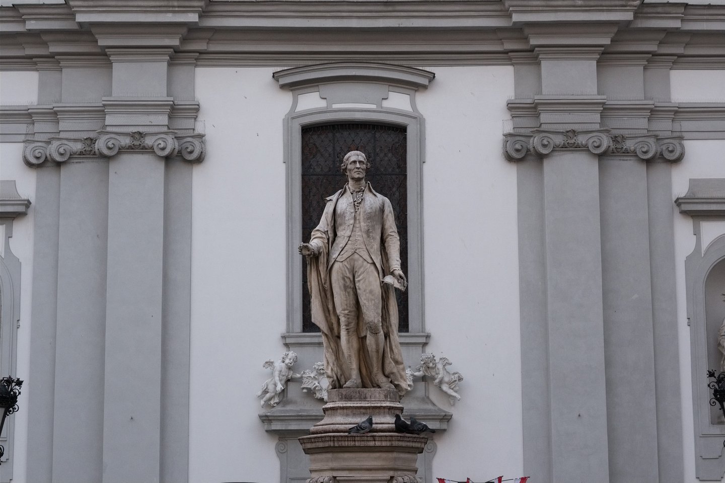 The stone statue of Joseph Haydn holding a quill and a sheet of music, standing on its pedestal in front of the pilastered facade of the Mariahilfer Kirche in Vienna.