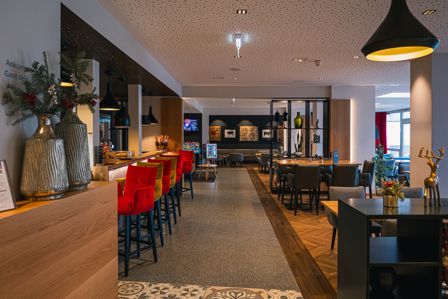 JUFA Hotel Salzburg, bar at Christmas, red and ochre velvet stools along a pale oak counter, a terrazzo aisle inlaid with a patterned tile panel and a dining room of dark chairs beyond.