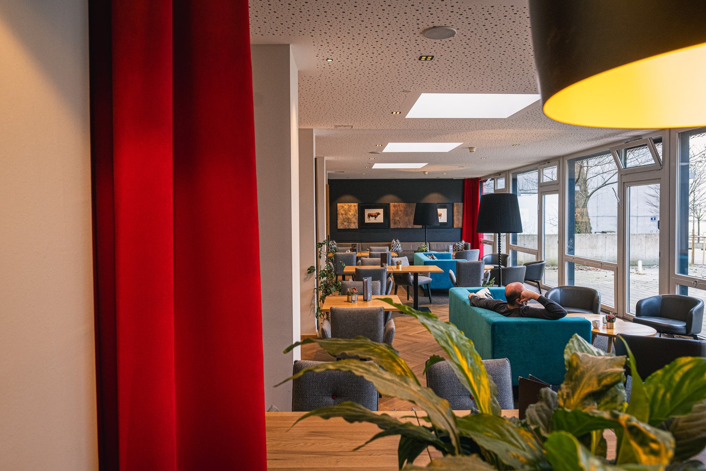 JUFA Hotel Salzburg, lounge seen past a floor-length red curtain, teal velvet sofas and grey tub chairs under pendant lamps, framed cattle prints on a dark wall and a glazed wall onto a winter terrace.