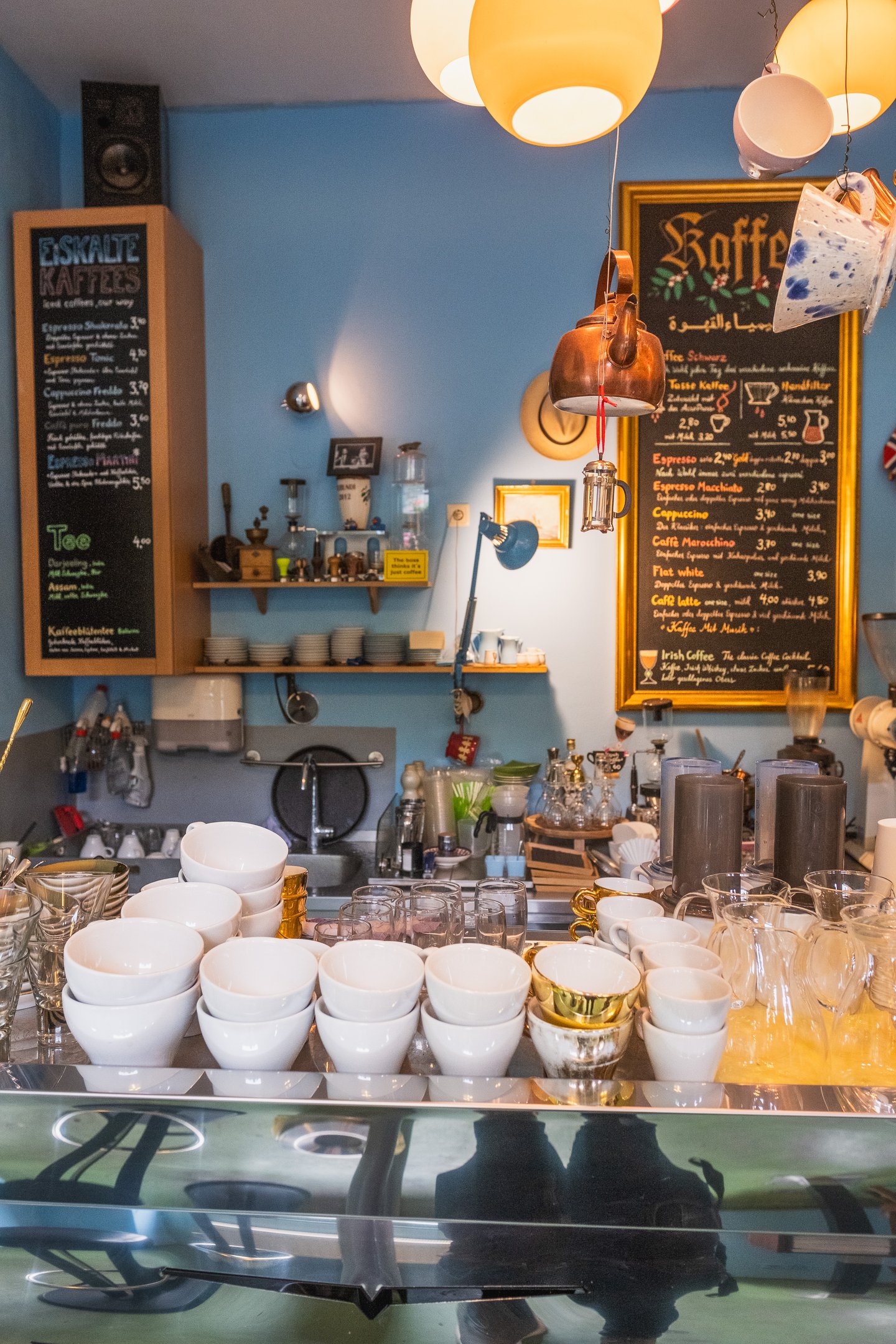 A vertical view across the counter of Kaffee Alchemie in Salzburg, ranks of white cups and glasses in the foreground and the full gold-framed coffee price board beside the Eiskalte Kaffees chalkboard on the blue wall behind.
