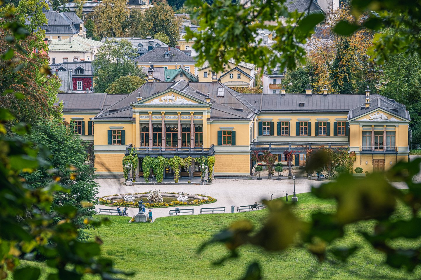 The full length of the Kaiservilla seen from a grassy slope through foliage, with the sculpted park fountain and benches on the gravel forecourt below it.