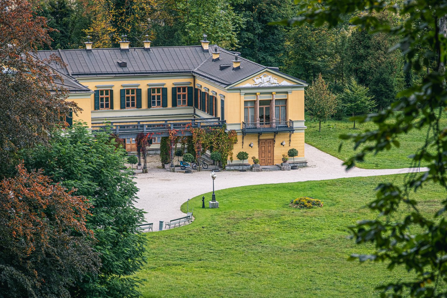 A side wing of the Kaiservilla viewed from higher in the park, its balcony hung with reddening creeper above the gravel drive and lawn.