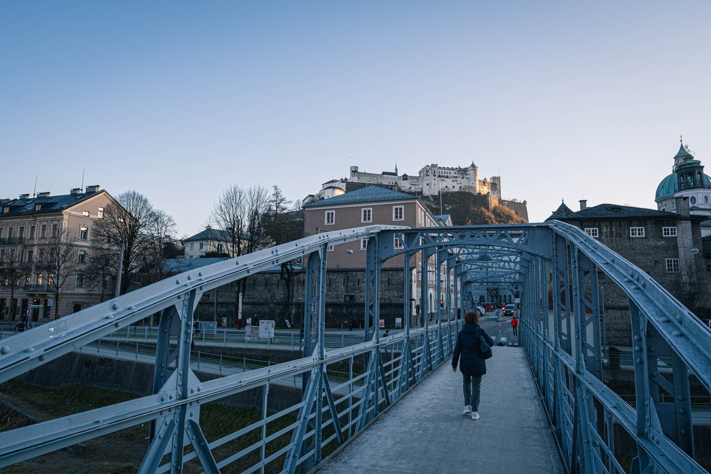 Pedestrian walking across the metal-arch Karolinenbruecke truss bridge with Hohensalzburg Fortress on the hill above the Altstadt in Salzburg