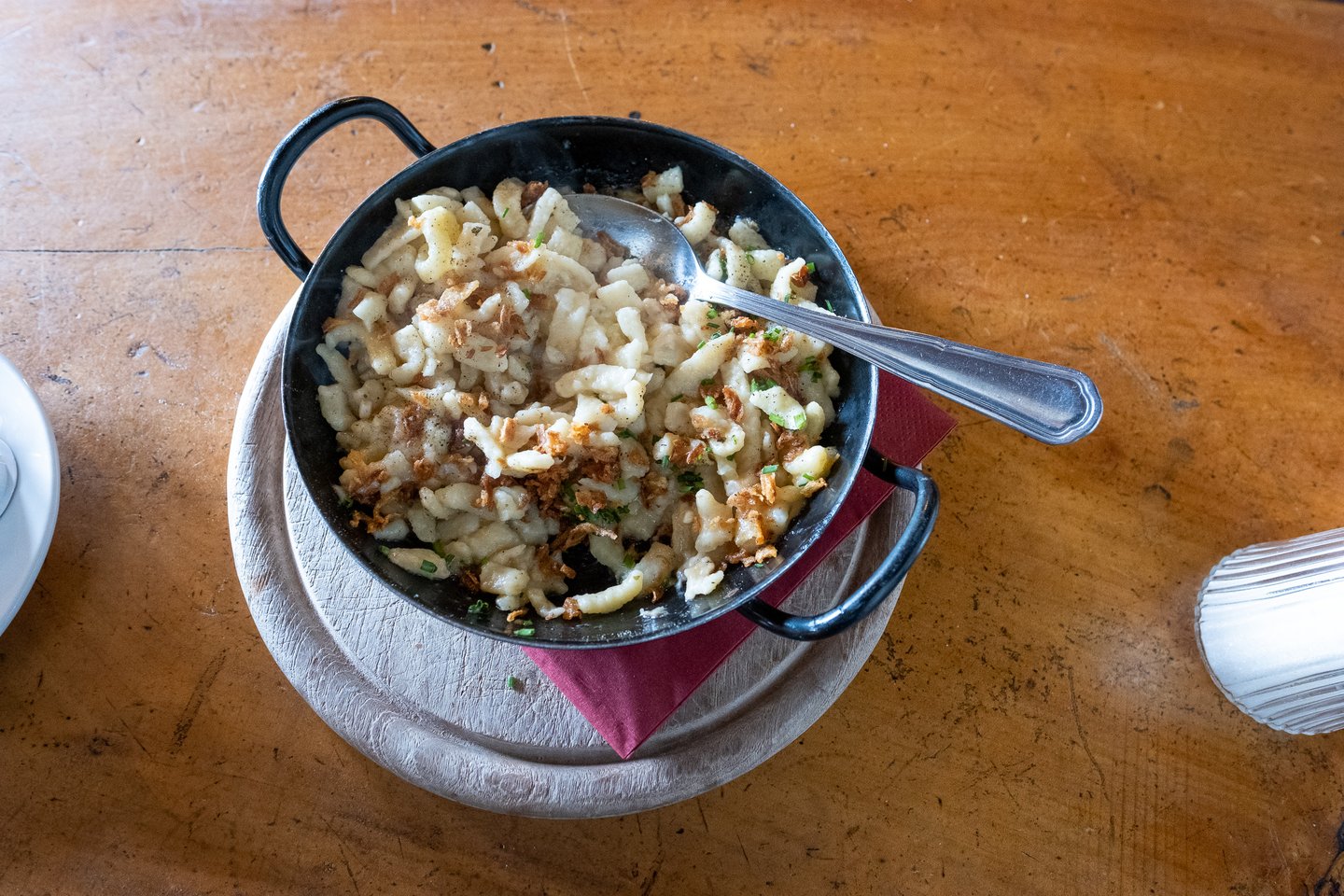 Overhead view of an iron pan of cheese Kasnocken with crisp fried onions and chives, resting on a pale wooden board on a scuffed wooden table.