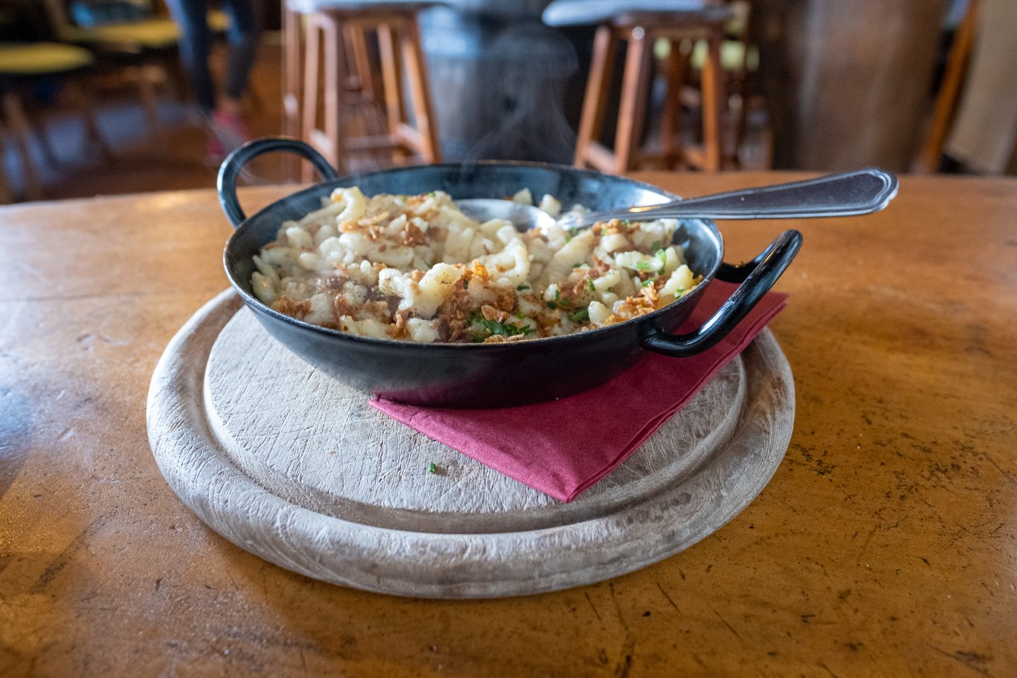 A steaming black iron pan of Kasnocken topped with crisp fried onions and chives sits on a pale wooden board, with the bar stools of a mountain hut behind.
