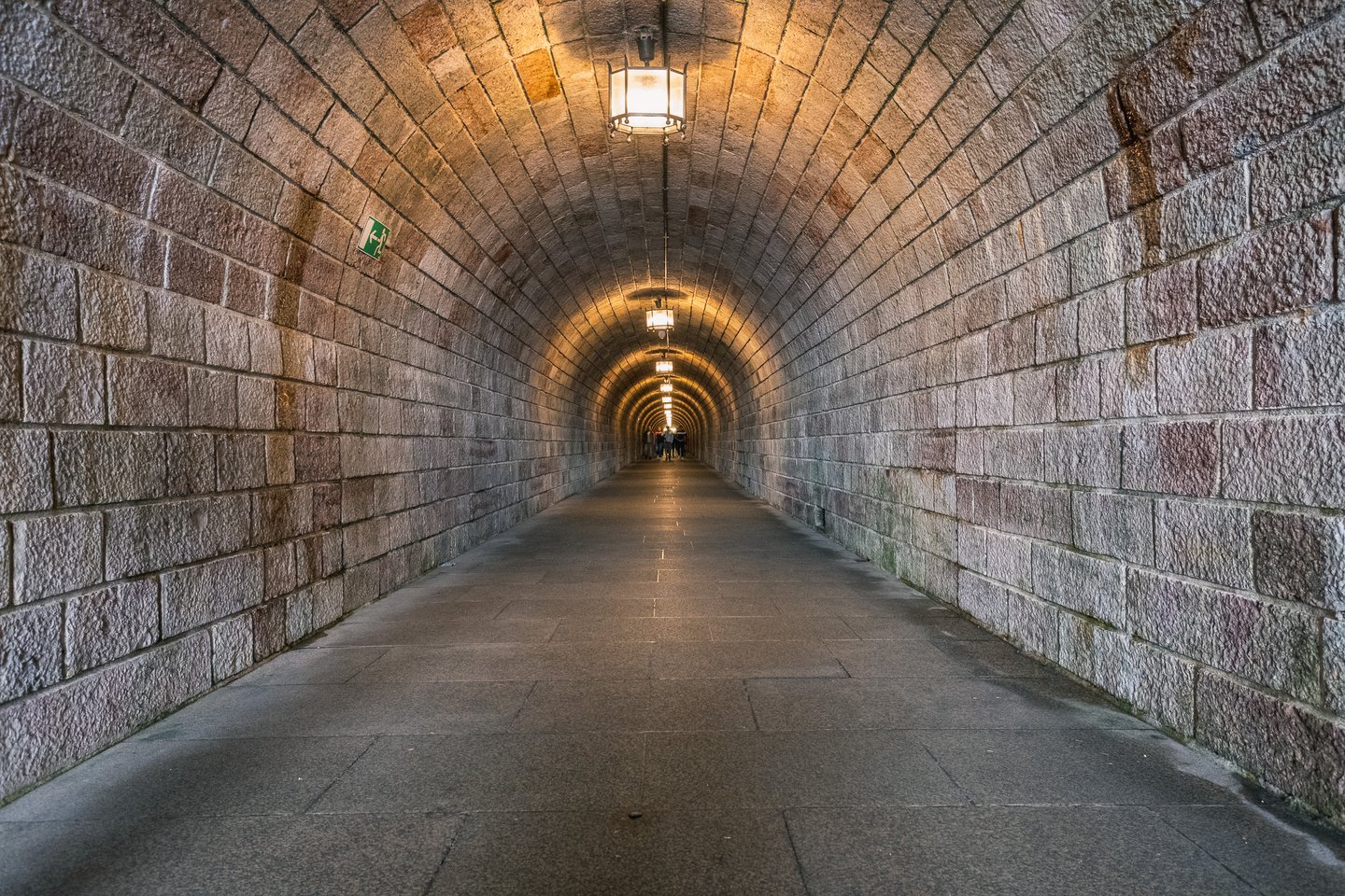 The long vaulted granite tunnel leading to the Kehlsteinhaus lift recedes into the distance, lit by hanging lanterns, with a few visitors at the far end.