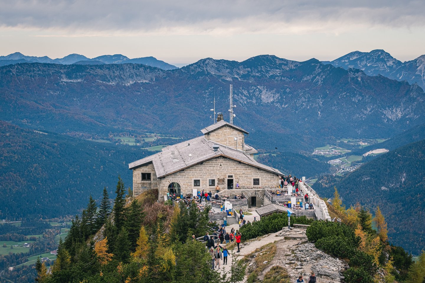 The Kehlsteinhaus sits on its rocky spur above the Berchtesgaden valley, its stone roof and aerial mast seen from the summit path with layered ranges behind.