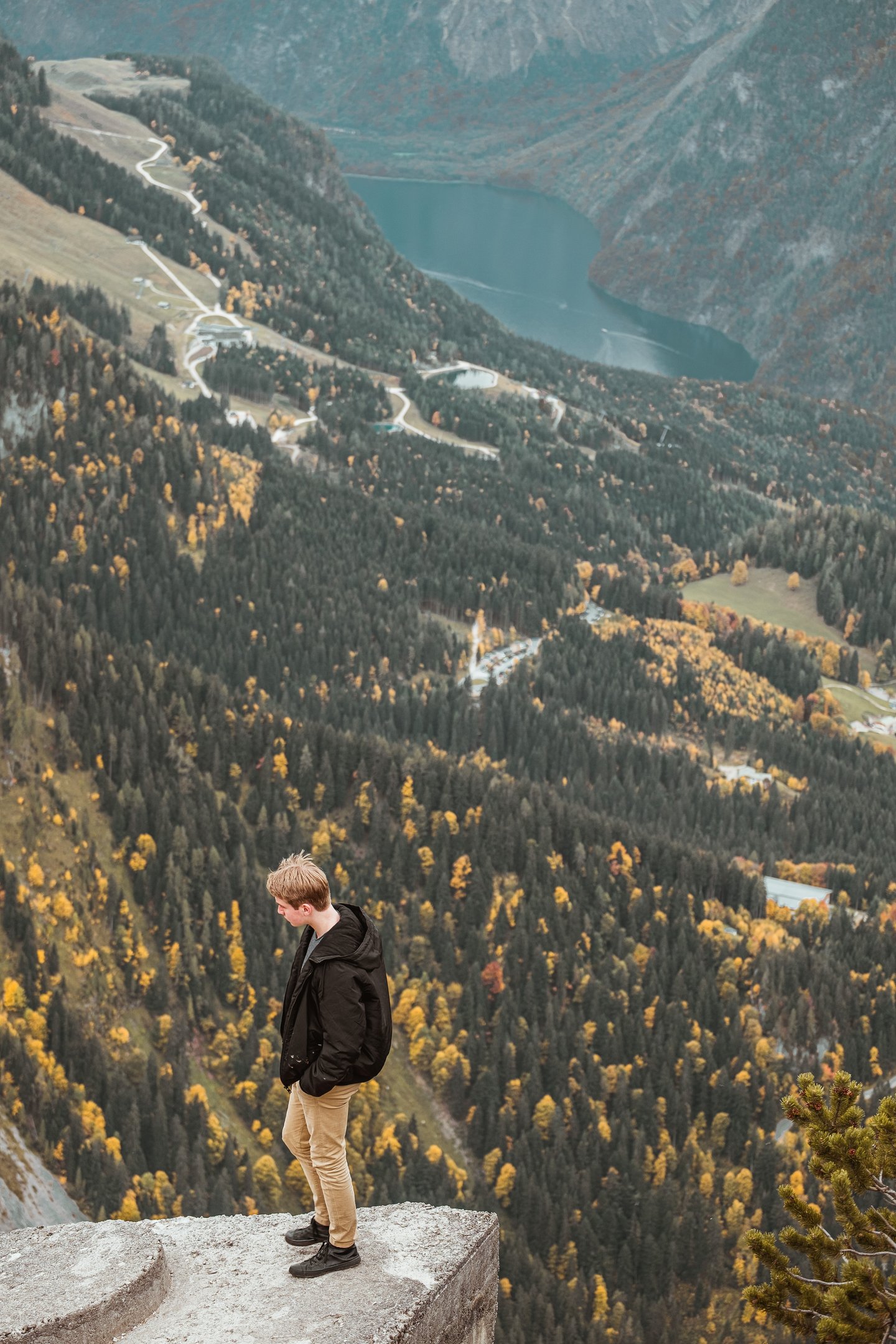 A young man stands on the edge of the Kehlstein concrete platform high above the Königssee, with the Jenner cable-car pistes zigzagging down the far slope.