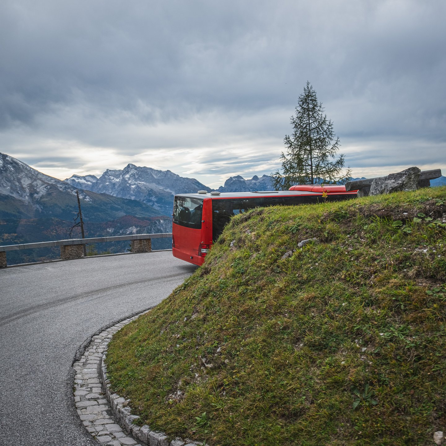 A red shuttle bus rounds a cobble-edged hairpin on the Kehlsteinstrasse, with a lone larch and the snow-dusted Berchtesgaden Alps beyond.