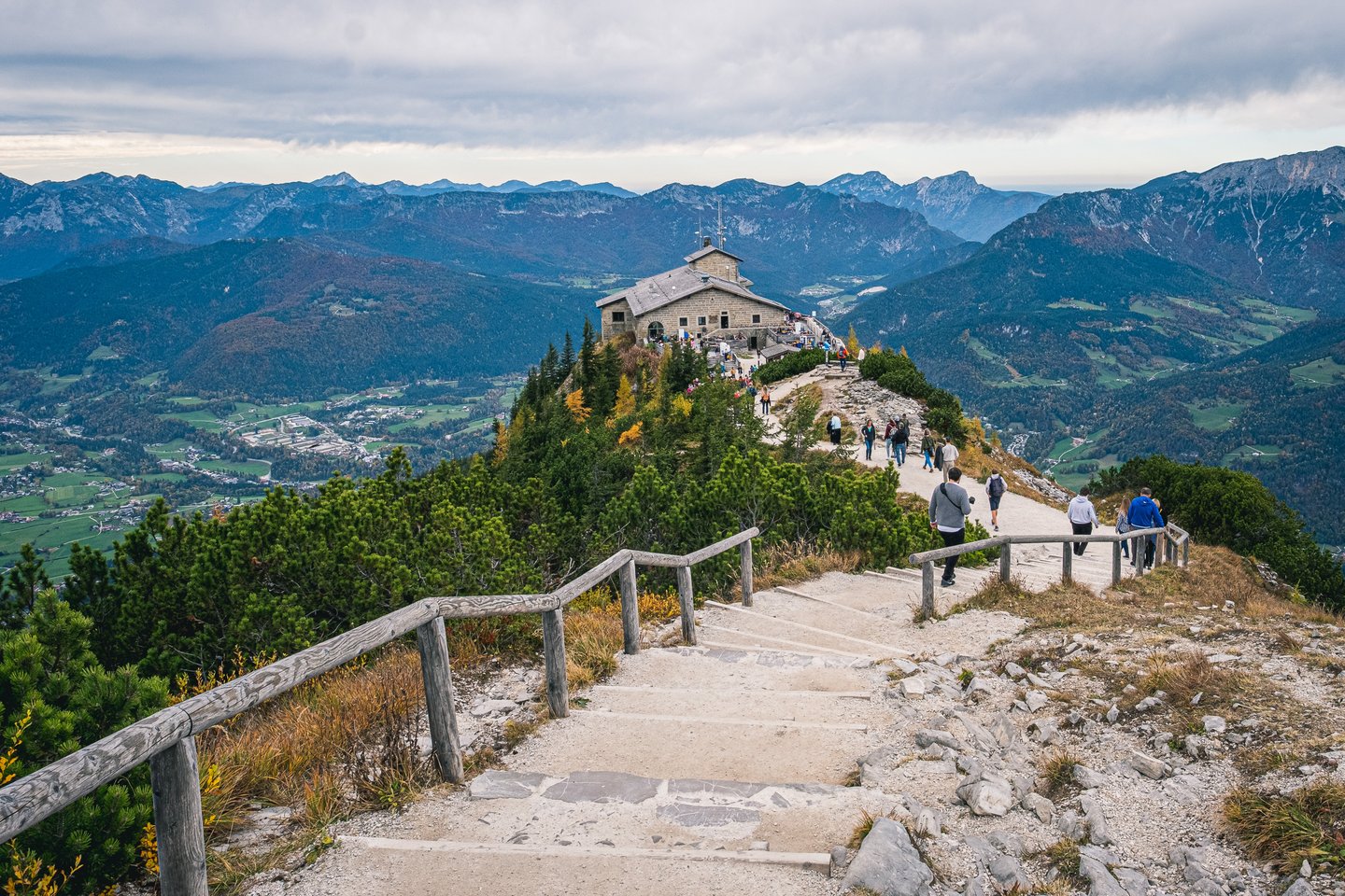 Stone steps with a weathered wooden handrail lead down the ridge from the Kehlstein summit to the Kehlsteinhaus, walkers strung out along the path.