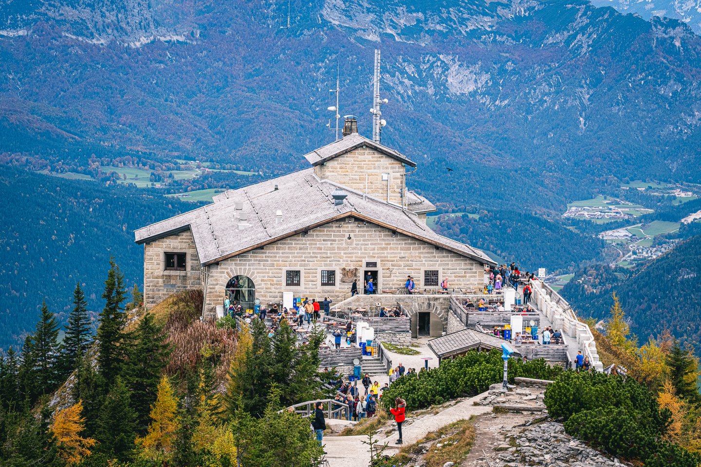 A closer view of the Kehlsteinhaus with visitors filling its stone terrace and benches, the Berchtesgaden valley and its dark ridges far below.