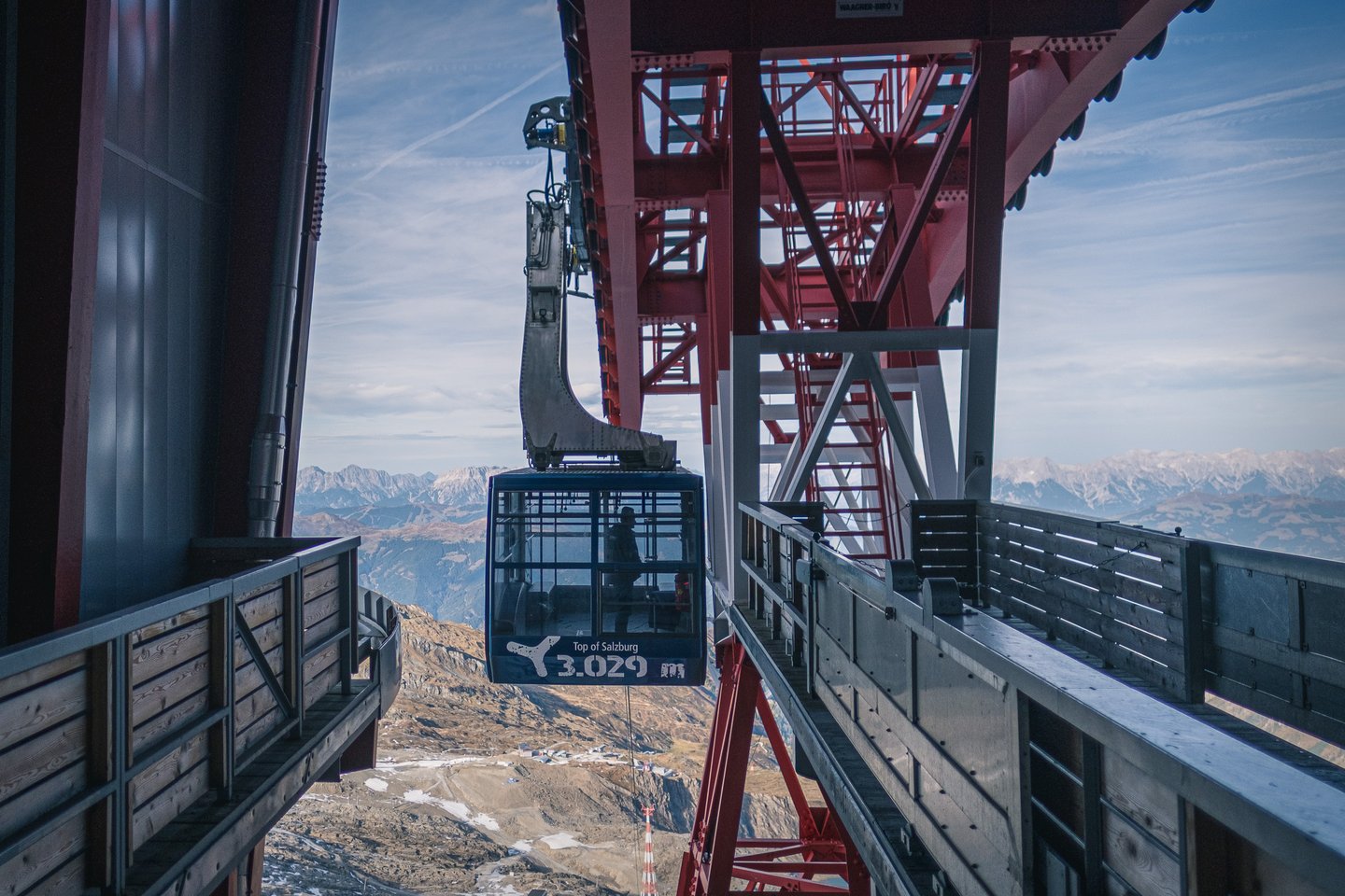 An aerial tram car lettered Top of Salzburg 3.029 m hangs from its red steel gantry as it leaves the Kitzsteinhorn summit station, with hazy ranges beyond.