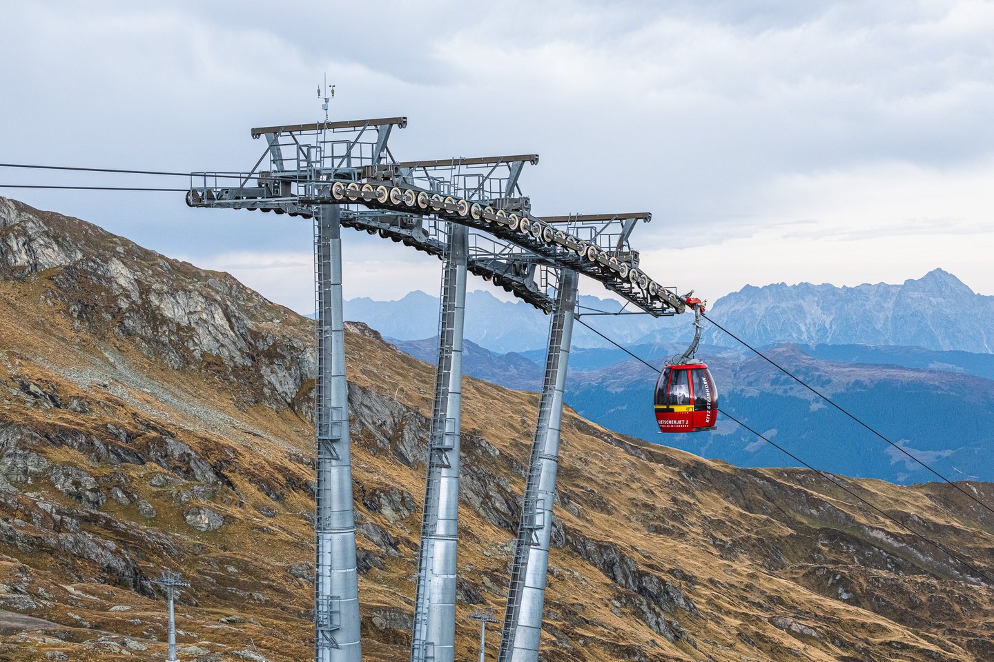 A red Gletscherjet 2 gondola of the Kitzsteinhorn lifts hangs between three tall steel pylons above brown autumn slopes, with limestone ranges fading into haze behind.