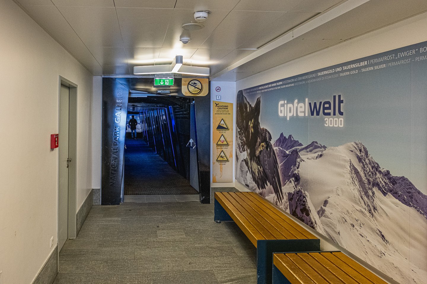 The lit corridor at the Kitzsteinhorn summit station leading into the dark Nationalpark Gallery tunnel, with the Gipfelwelt 3000 mural and its golden eagle running along the right wall.