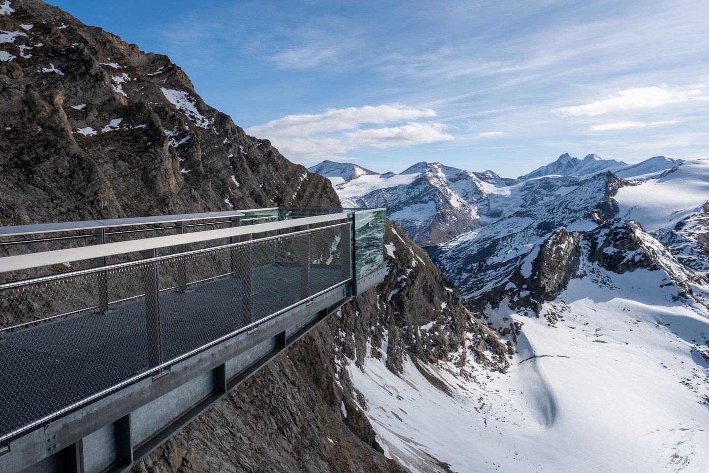 The steel skywalk gangway at the Kitzsteinhorn summit runs out along the cliff edge, with glaciated Hohe Tauern peaks and one sharp dark summit on the horizon beyond.