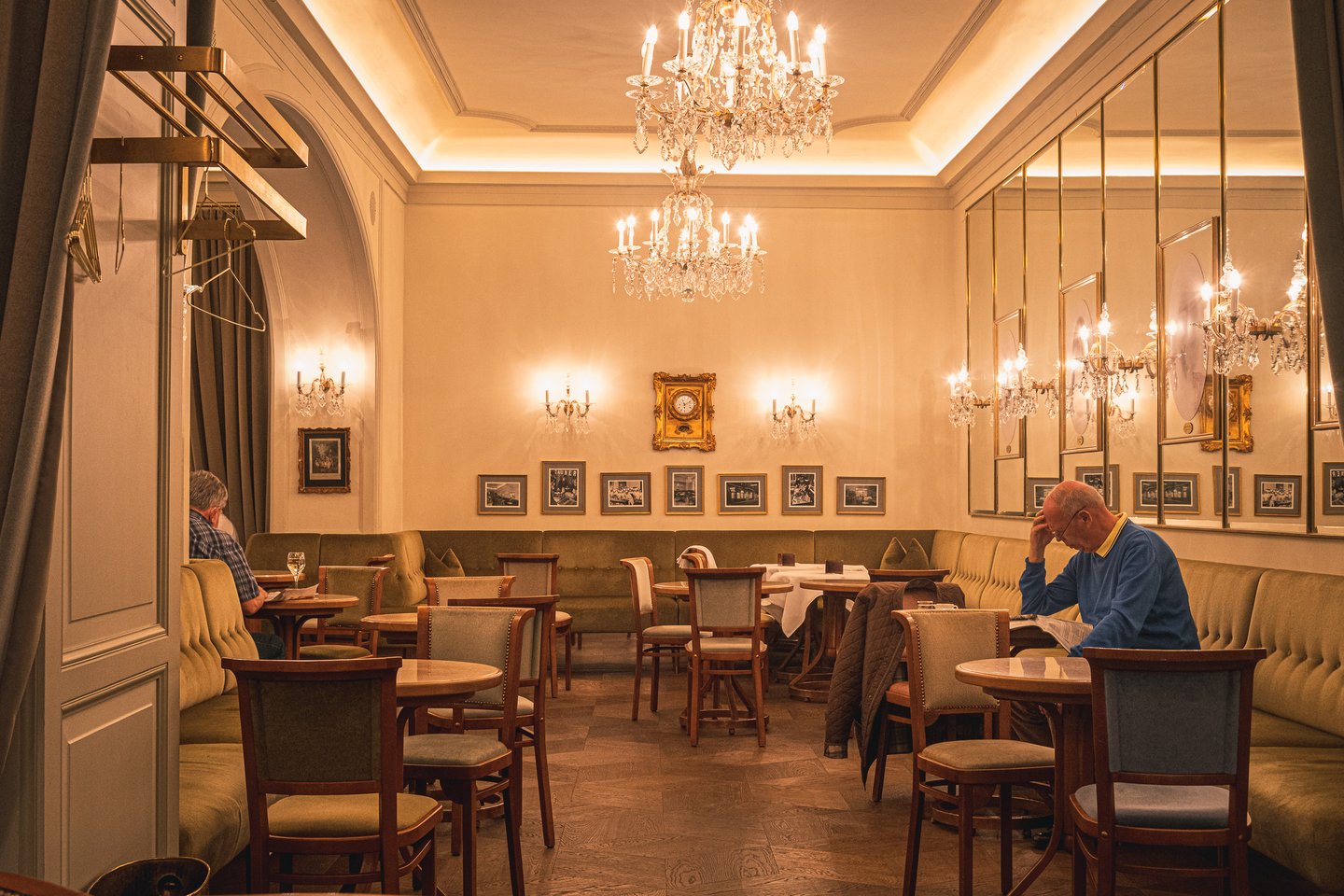 The warmly lit cafe salon at Zauner in Bad Ischl, with crystal chandeliers, olive banquettes, bentwood chairs and two guests reading at marble tables.