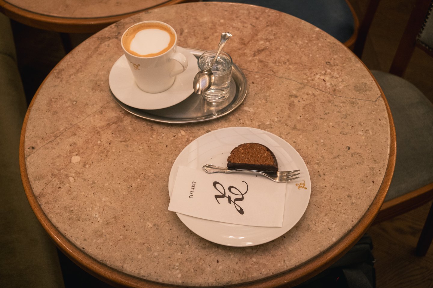 Seen from above, a melange on a silver tray with a water glass beside a slice of chocolate-glazed cake on a Zauner plate, on a round marble cafe table.