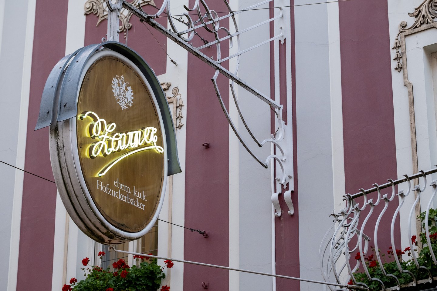 The oval Zauner sign hanging from an ornate iron bracket, its script name in lit neon under a double-headed eagle and the line ehem. k.u.k. Hofzuckerbaecker.
