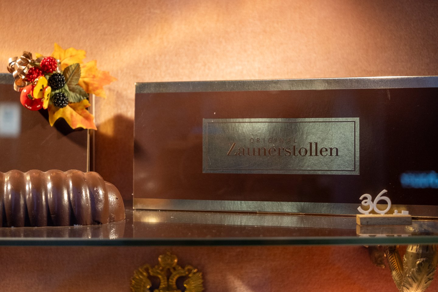Close view of a brown presentation box in the Zauner window, its gold panel reading Original Zaunerstollen, with the end of the cake and an autumn decoration beside it.