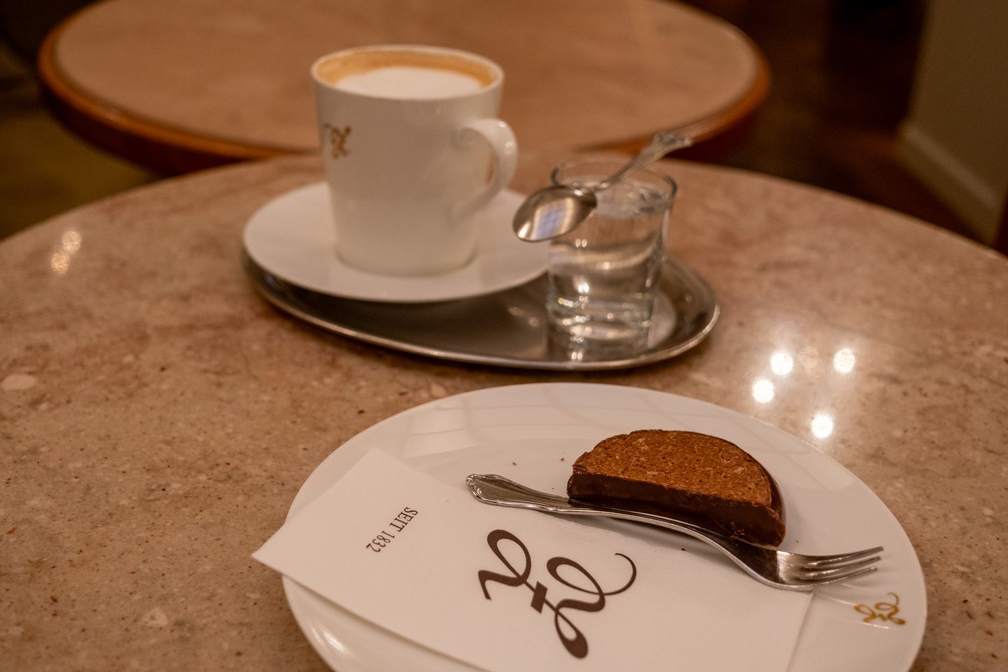 A low view across the marble table at Zauner, with the chocolate-coated cake slice on its monogrammed napkin in front and the melange on its silver tray behind.