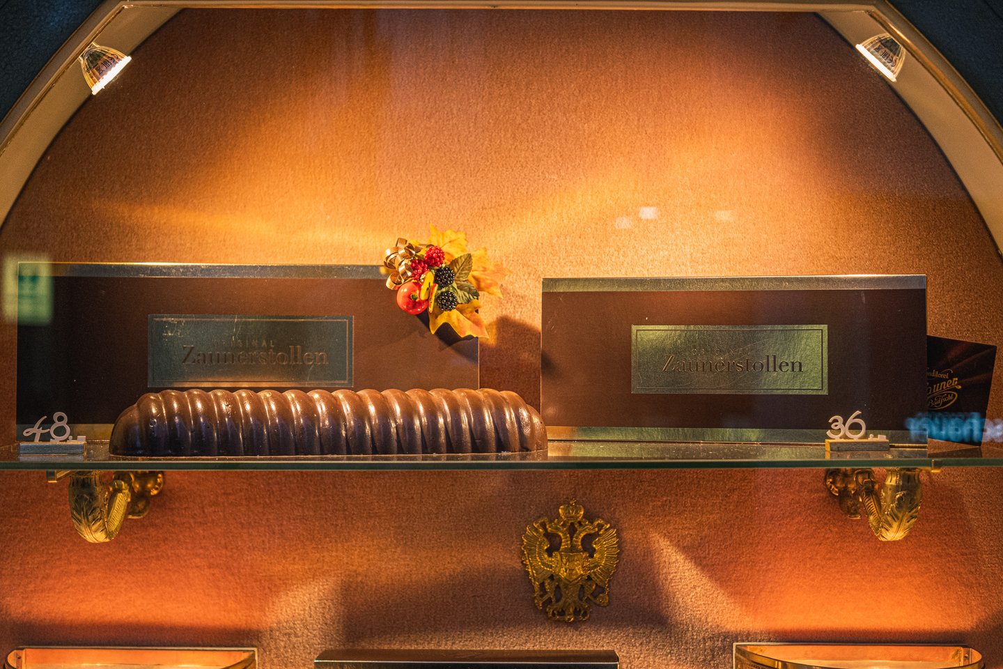 A glass shelf in the Zauner window holding a long ribbed Zaunerstollen beside two brown presentation boxes, lit warm above a gilded double-headed eagle.