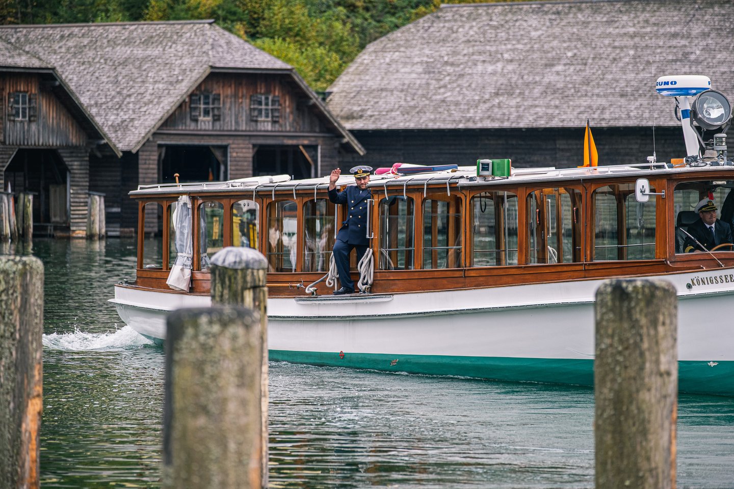 The captain of a Koenigssee tour boat waves from the open foredeck in cap and uniform as the vessel passes a row of old shingled wooden boathouses.