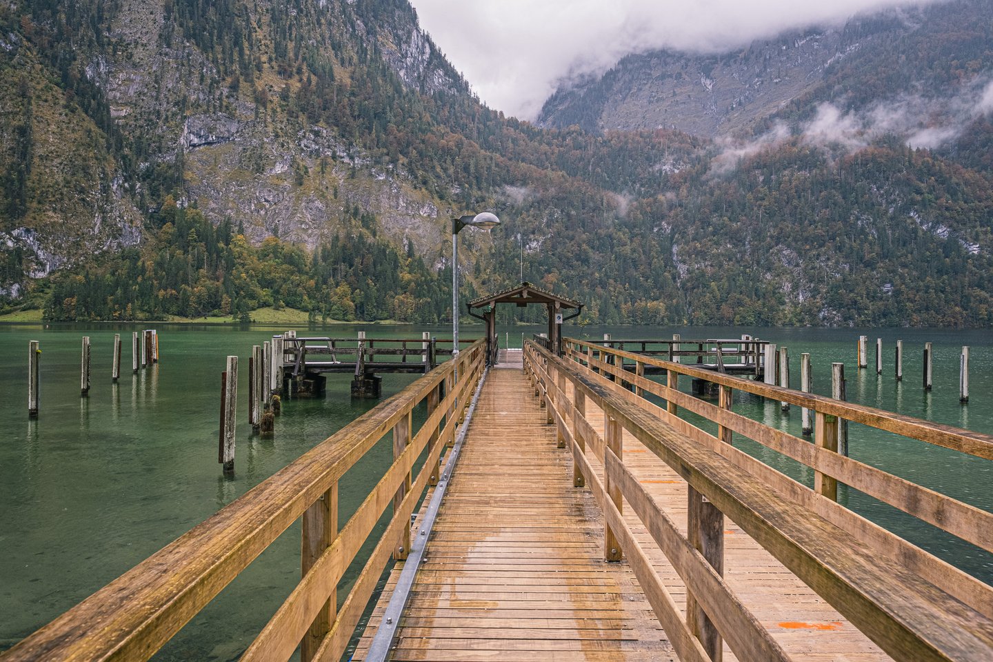 The long empty timber boardwalk of the Salet landing runs out over the green water of the Koenigssee towards a small shingled shelter, with cloud on the cliffs beyond.
