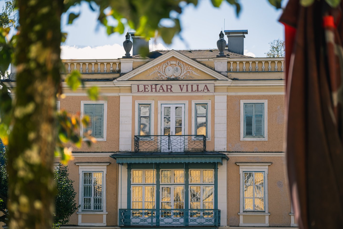 The ochre facade of the Lehar Villa in Bad Ischl, its name painted in red under the pediment, glimpsed between foreground foliage.
