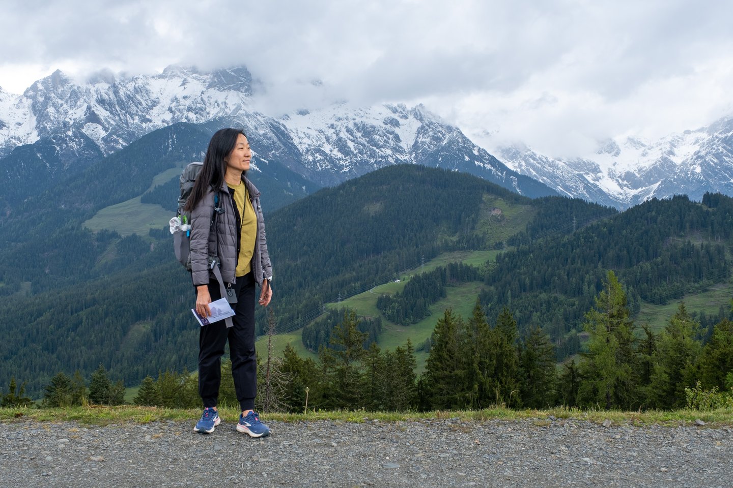 A hiker stands in profile holding a folded leaflet and looks out across the wooded Urslau valley toward the snow-covered range above Maria Alm.