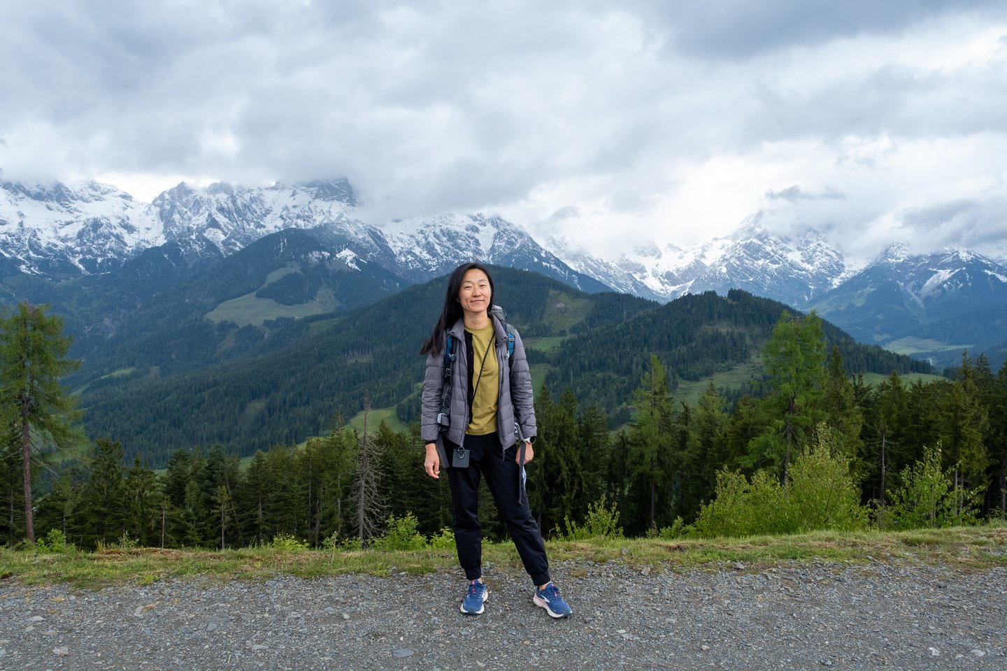 A woman in a grey down jacket stands centred on a gravel track high on the Natrun above Maria Alm, with the snow-covered limestone wall of the Steinernes Meer rising behind forested slopes.